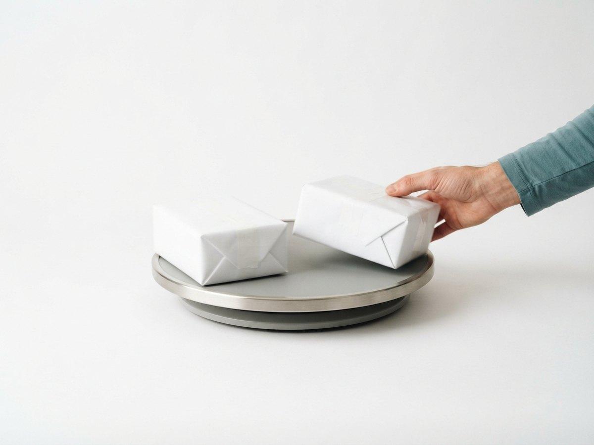 A hand places one of two white wrapped parcels onto a round metal and gray tray against a plain white background