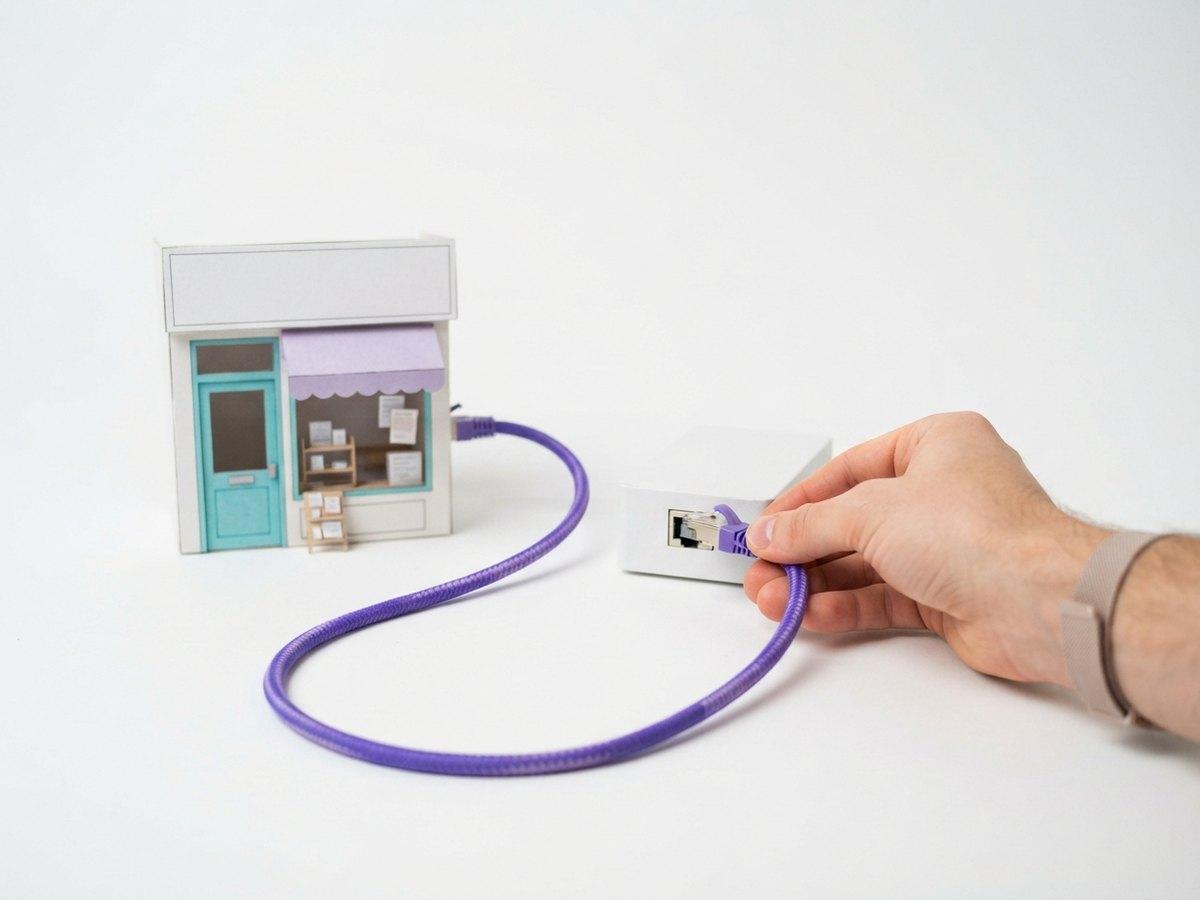 A hand plugs a purple network cable into a white box, its other end attached to a small paper model shop