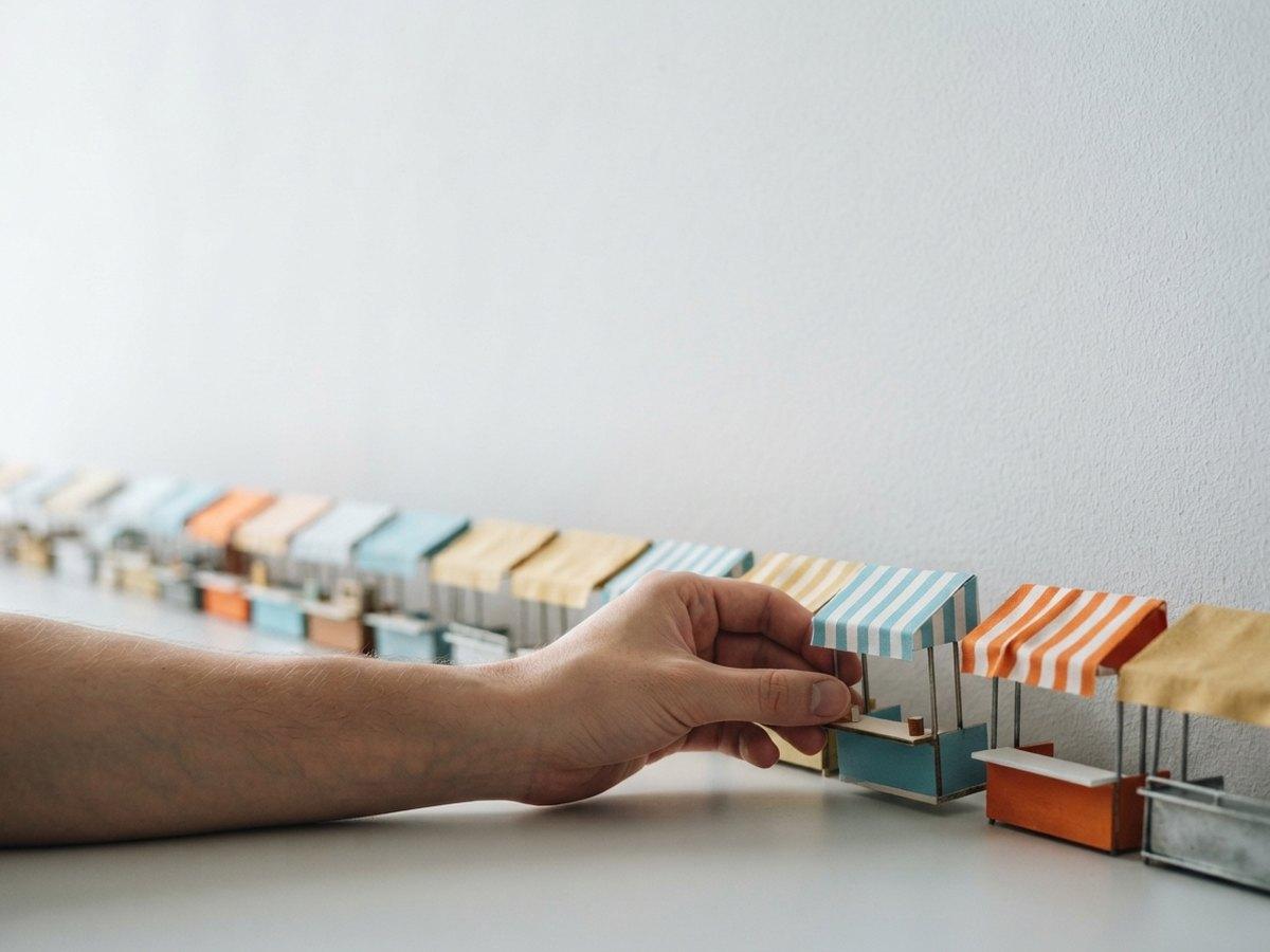 A hand adjusts a miniature striped market stall at the head of a long row of tiny stalls receding along a wall