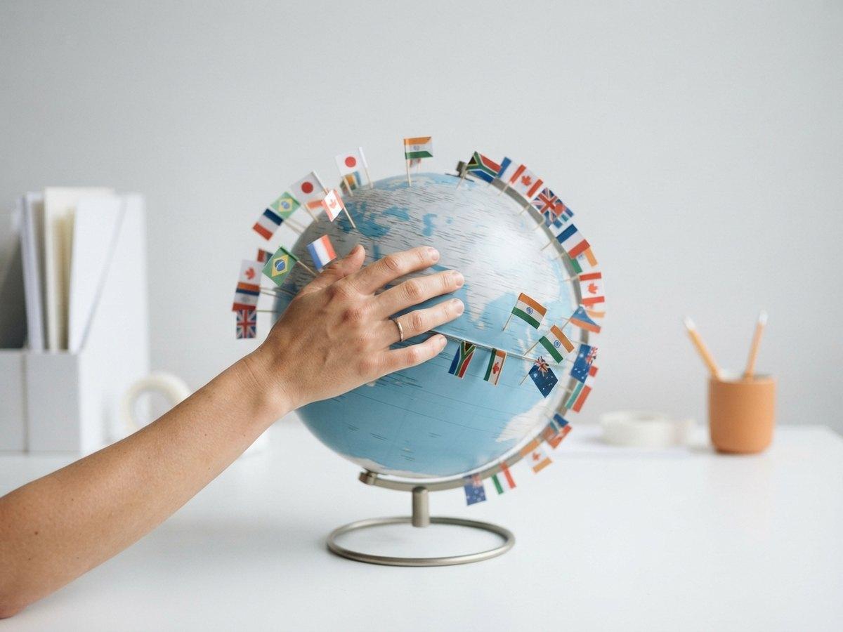 A hand rests on a blue desk globe studded with small national flags, with a pot of pencils behind