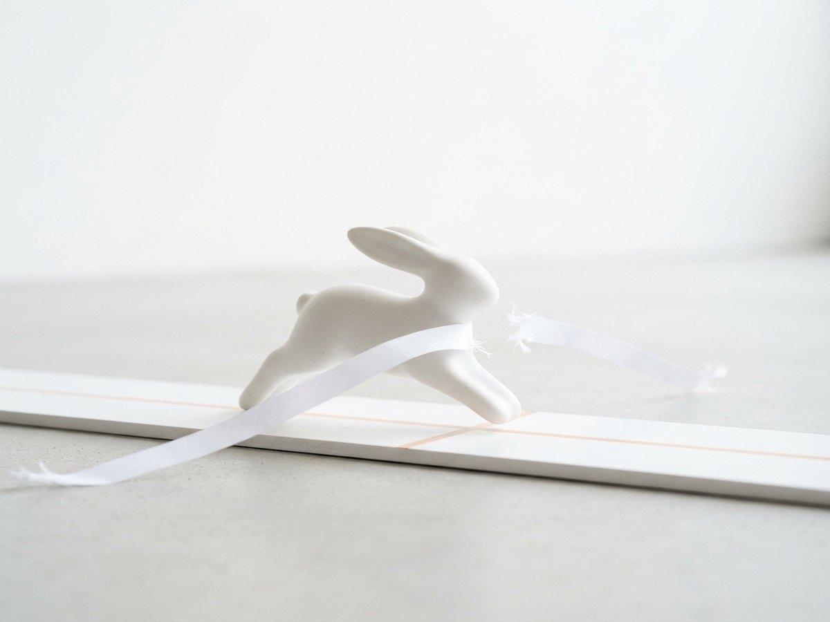A white porcelain hare leaps through a thin paper ribbon stretched across a white track like a finish line