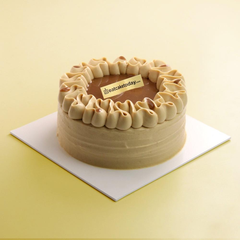 Pandan gula melaka cake with ruffled caramel-colored cream around a caramel top and a small gold eatcaketoday.com label, on a white board against a pale yellow background
