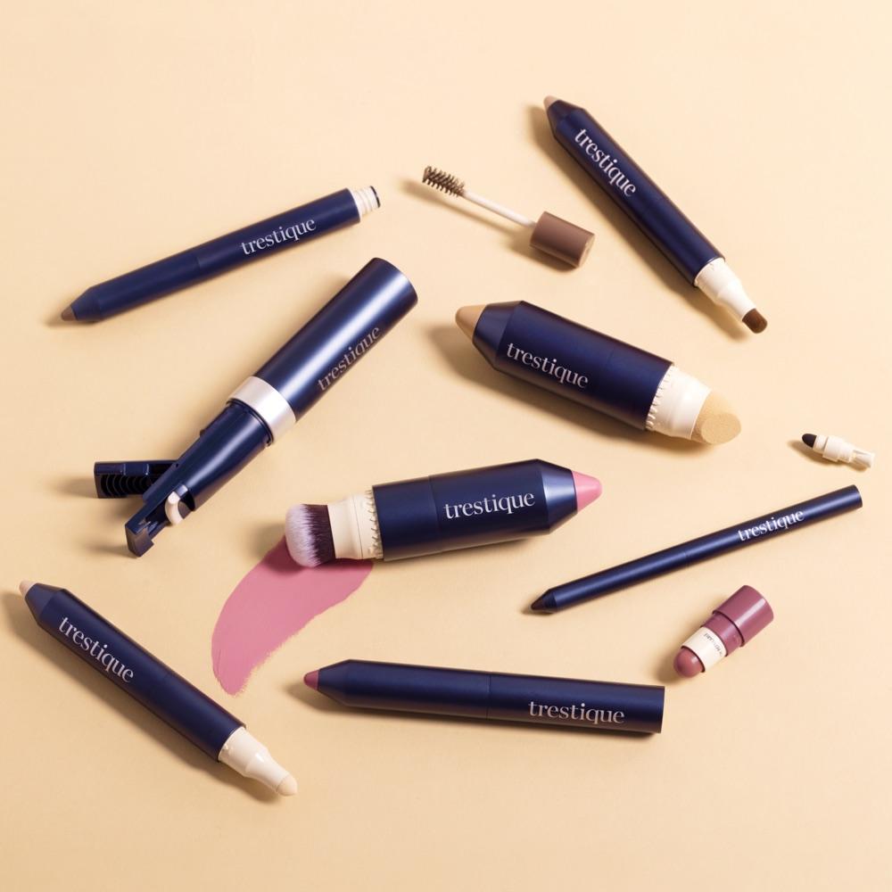 Flat lay of eight Trestique navy and cream makeup pieces, including a blush stick with a brush and a foundation stick with a sponge, on a warm cream background