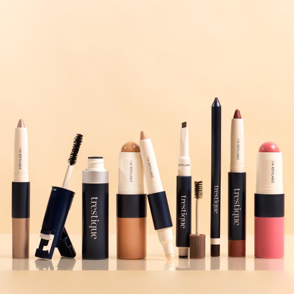 Row of Trestique makeup pieces, including a mascara, a foundation stick, a concealer crayon, a brow pencil and a blush stick, in front of a cream backdrop
