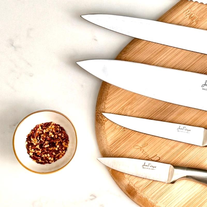 Jean Patrique Master Gourmet knife blades on a round bamboo board beside a small bowl of chili flakes on a marble surface